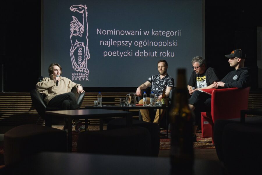 Czterech mężczyzn siedzi za stołem. Z tyłu wyświetlony jest napis: "Nominowani w kategorii najlepszy ogólnopolski poetycki debiut roku"