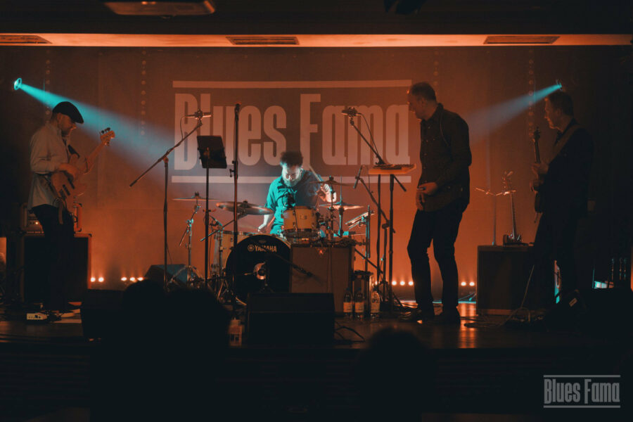 4 osoby na scenie, basista, perkusista, wokalista i gitarzysta. Z tyłu napis "Blues Fama".