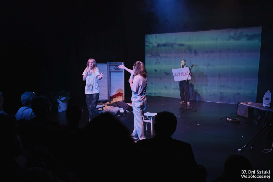 Troje osób na scenie, dwie kobiety i mężczyzna z kartką z napisem "APLAUZ", otwarta lodówka, porozrzucane ubrania