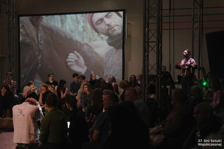 Publiczność, wśród ludzi stoi aktor, z tyłu wyświetlane zdjęcie człowieka z koniem