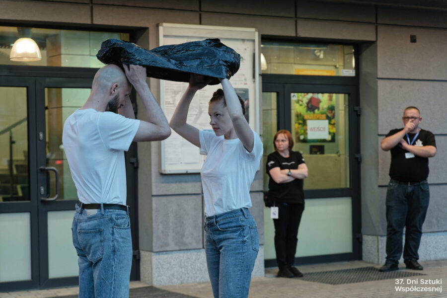 Dwoje performerów wykonuje performans "Stretching" przed budynkiem BOK-u, trzymają folię nad głową.