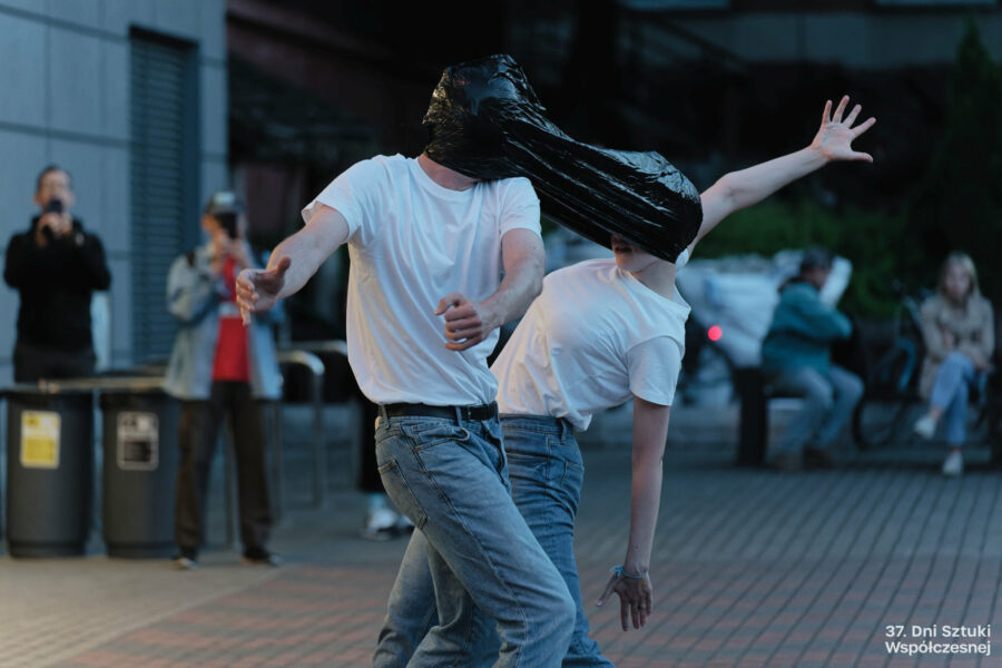 Dwoje performerów wykonuje performans "Stretching" przed budynkiem BOK-u, mają głowy związane folią.