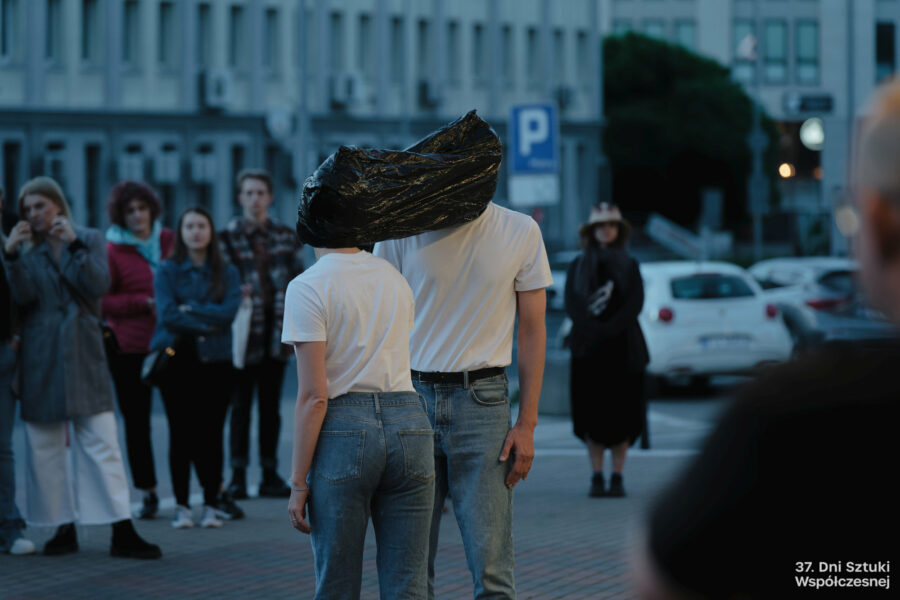 Dwoje performerów wykonuje performans "Stretching" przed budynkiem BOK-u, mają głowy związane folią. W tle publiczność.